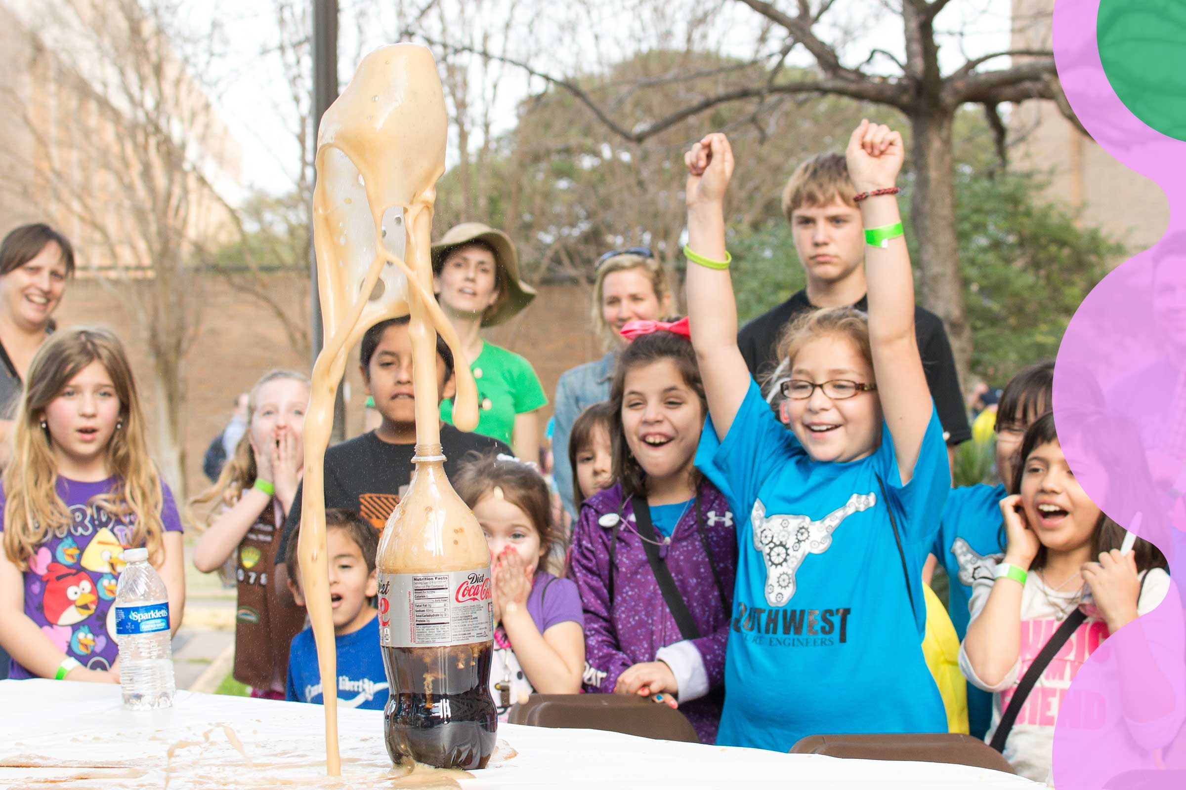 About a dozen children cheer as a fountain of soda shoots up out of a soda bottle in a scientific demo.