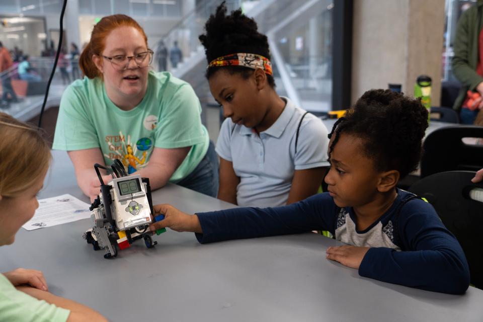Volunteer college students in STEM Girl Day shirts demo a device of legos with robotic overtones to a pair of school-aged girls.